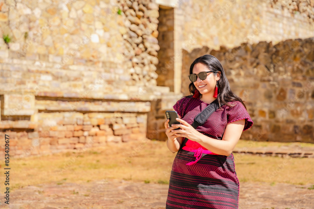 Fototapeta premium Joyful young Latina woman in traditional dress takes a selfie at Monte Alban ruins in Oaxaca, Mexico, capturing her adventure with smartphone.