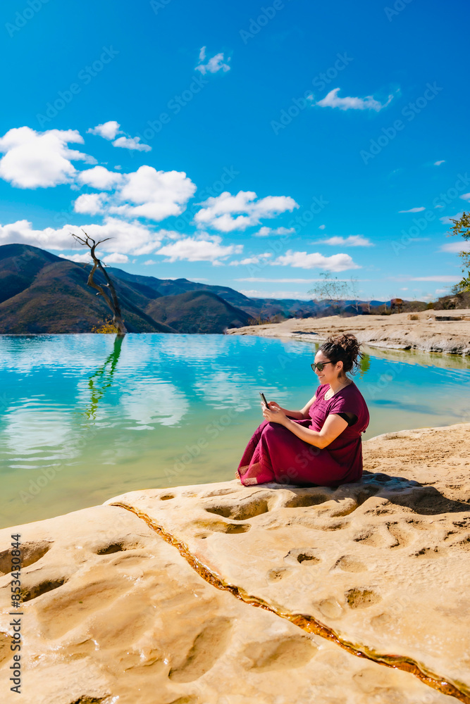 Obraz premium Young relaxed Latina woman in her twenties, dressed in traditional clothing, sits by the tranquil waters of Hierve el Agua in Oaxaca, Mexico, using her smartphone.