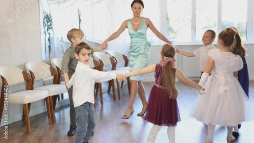 Happy little children in elegant dresses doing round dance with their teacher in school hall  photo