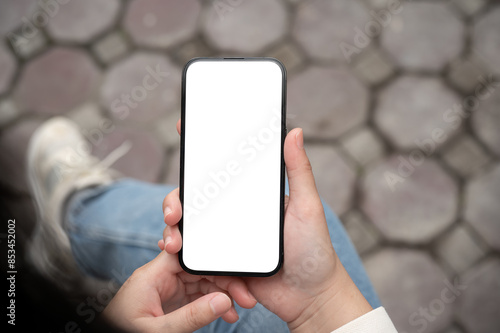 A woman holding a smartphone with a white-screen mockup over her lap while sitting outdoors.