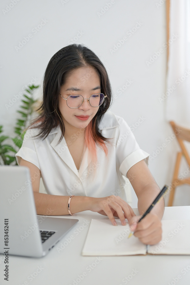 A focused Asian woman is working remotely from a cafe, taking notes or making list in her notebook A focused Asian woman is working remotely from a cafe, taking notes or making list in her notebook