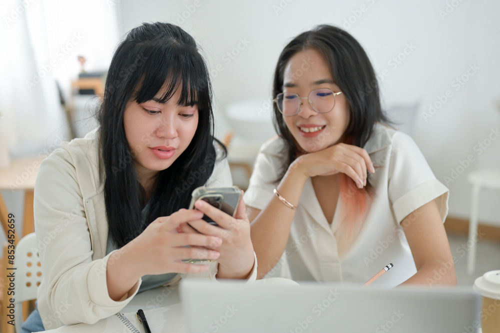Two Asian women are sitting in a cafe, talking while looking at a smartphone screen together. Two Asian women are sitting in a cafe, talking while looking at a smartphone screen together.