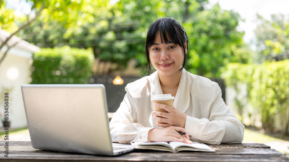 A charming young Asian businesswoman sits at a wooden table in a garden, smiling at the camera. A charming young Asian businesswoman sits at a wooden table in a garden, smiling at the camera.