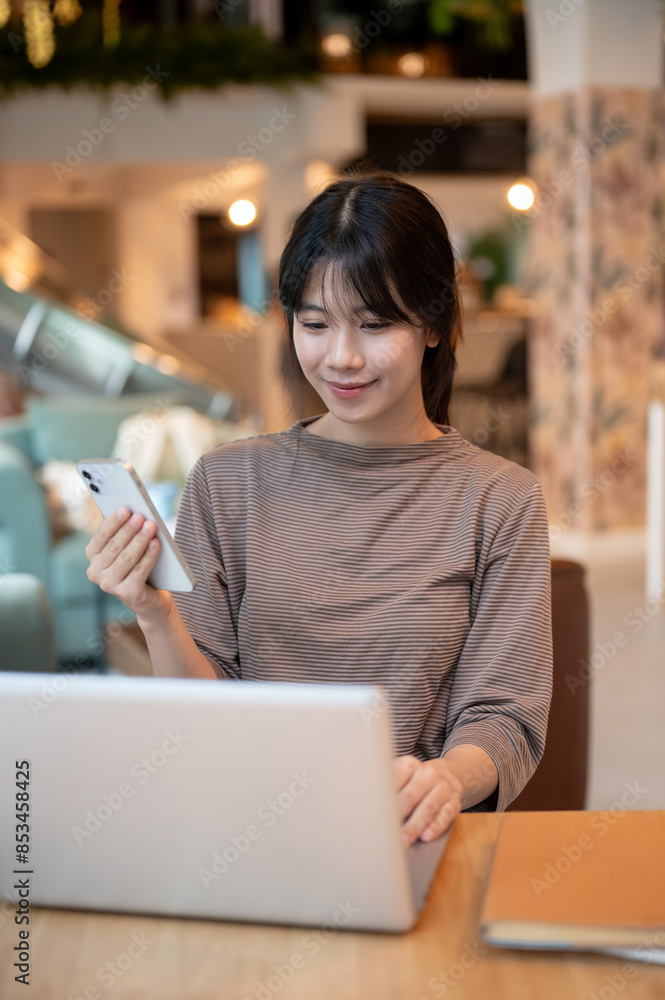 A beautiful Asian woman is holding her smartphone while working on her laptop computer. A beautiful Asian woman is holding her smartphone while working on her laptop computer.