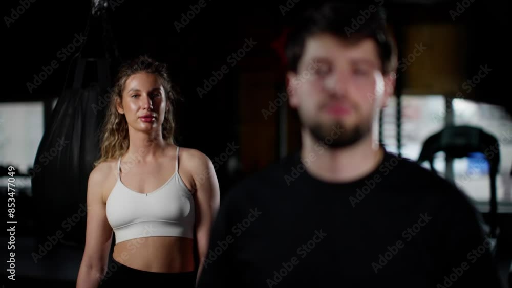 Focused man in black shirt and determined woman in white sports bra with boxing gloves in a dimly lit gym, standing in front of a punching bag, preparing for a boxing workout. Focused man in black shirt and determined woman in white sports bra with boxing gloves in a dimly lit gym, standing in front of a punching bag, preparing for a boxing workout.