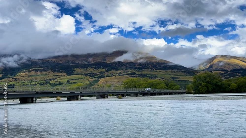 A single car drives across the Reese River Bridge on Glenorchy Paradise Road in New Zealand in front of large hills on a cloudy day photo