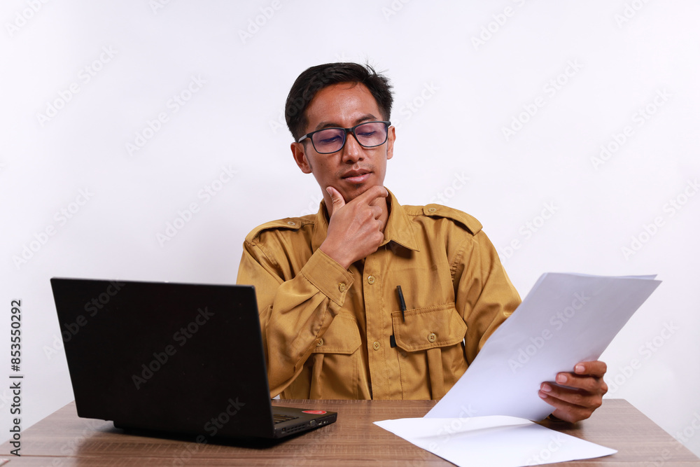 Male teacher in civil servant uniform with paper and laptop on the desk thinking something Male teacher in civil servant uniform with paper and laptop on the desk thinking something