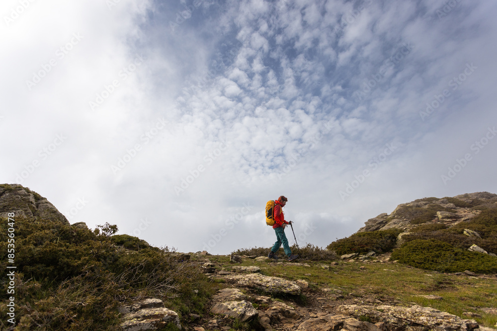 A woman in a red jacket is walking on a rocky hillside. The sky is cloudy, and the man is carrying a backpack. Concept of adventure and exploration A woman in a red jacket is walking on a rocky hillside. The sky is cloudy, and the man is carrying a backpack. Concept of adventure and exploration
