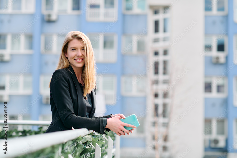Fototapeta premium Blonde Businesswoman in Black Suit Using Smartphone on Break in Modern Urban Environment.