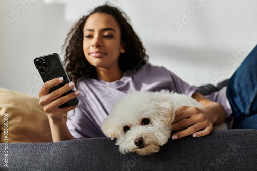 Photography A woman relaxes on a couch, holding her bichon frise dog and a cell phone