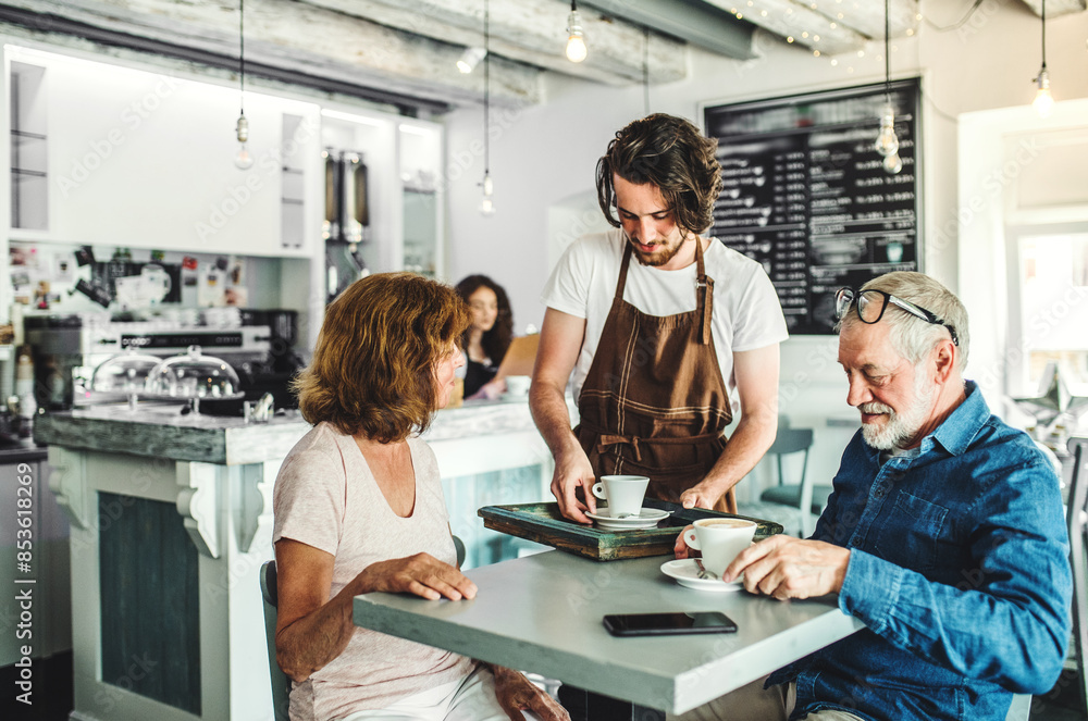 Fototapeta premium Handsome barista working in coffee shop, serving customers, elderly couple. University student working part-time in cafe.