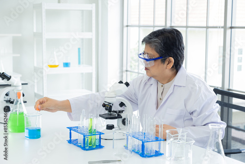 Professional senior scientist woman wearing safety goggles and a white lab coat working with a microscope in a modern laboratory. Various lab equipment and chemical solutions are shown on the table
