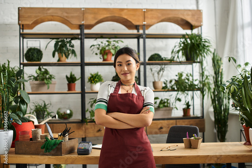 Obraz na plátně A beautiful Asian woman stands proudly in her plant shop, arms crossed, wearing an apron