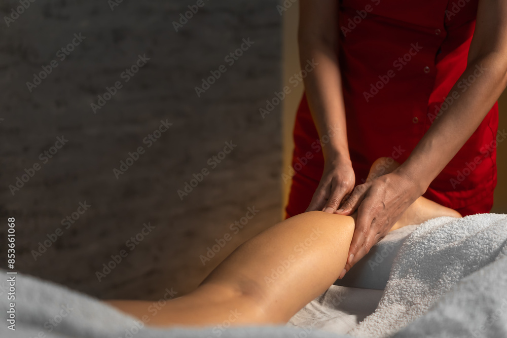 Fototapeta premium Close-up of a masseur in a red suit massaging the calf muscle of a young woman. Spa massage in a cozy massage therapist's office