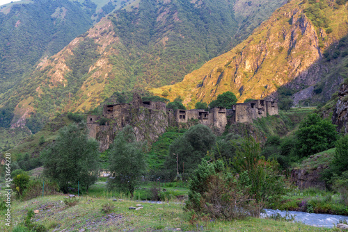 The morning sun illuminates the rock behind the medieval village of Shatili. Bushes and river in the foreground photo