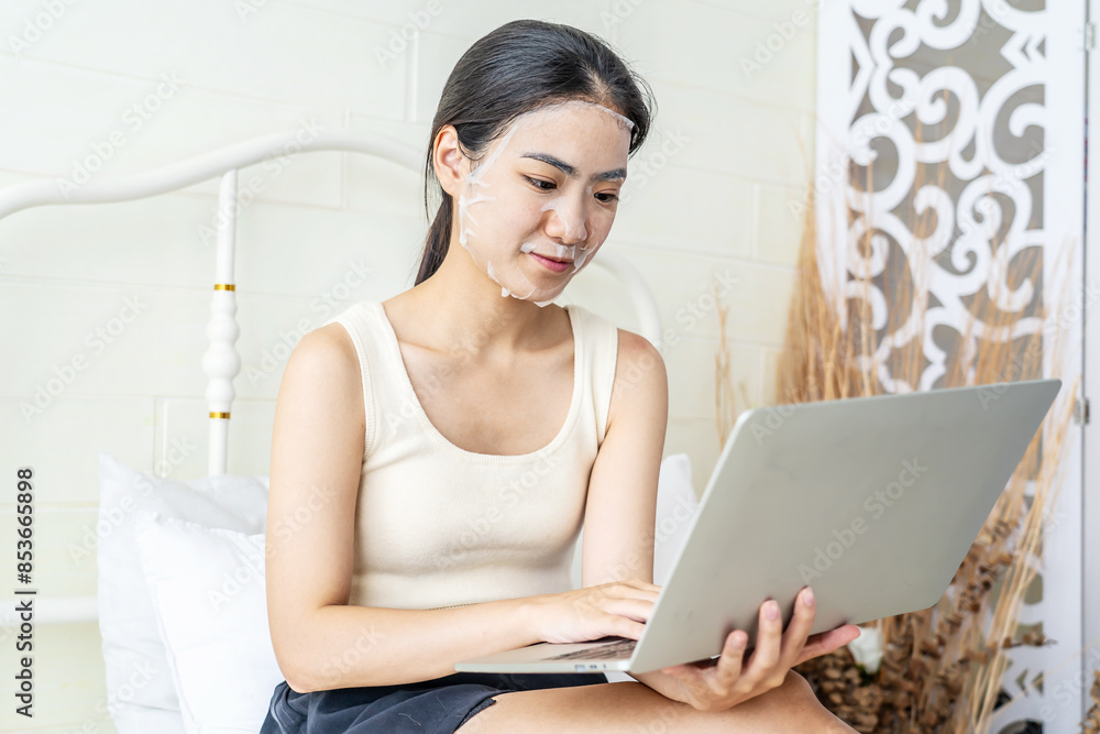 Relaxed beautiful young asian woman with sheet facial mask lying working on laptop computer on bed, beauty girl applying face mask for skincare of wrinkle at home, skin care, health and wellness. Relaxed beautiful young asian woman with sheet facial mask lying working on laptop computer on bed, beauty girl applying face mask for skincare of wrinkle at home, skin care, health and wellness.
