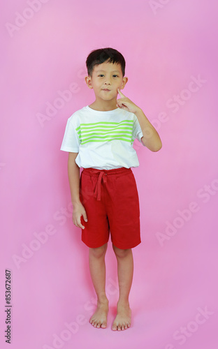 Cute Asian little boy kid age 7 years old keeping fingers on cheek. Child standing and looking straight at camera isolated on pink studio background. Image full length. © zilvergolf