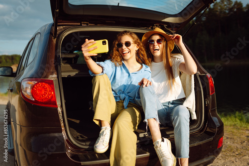 Young woman sits in the trunk of a car and takes selfie. Stylish models on a car trip. The concept of technology, blogging, travel. © maxbelchenko