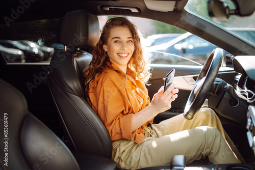 Beautiful young woman sitting in a car on drivers seat and using smartphone. Car sharing, rental service or taxi app. Lifestyle, travel, tourism, active life. photo