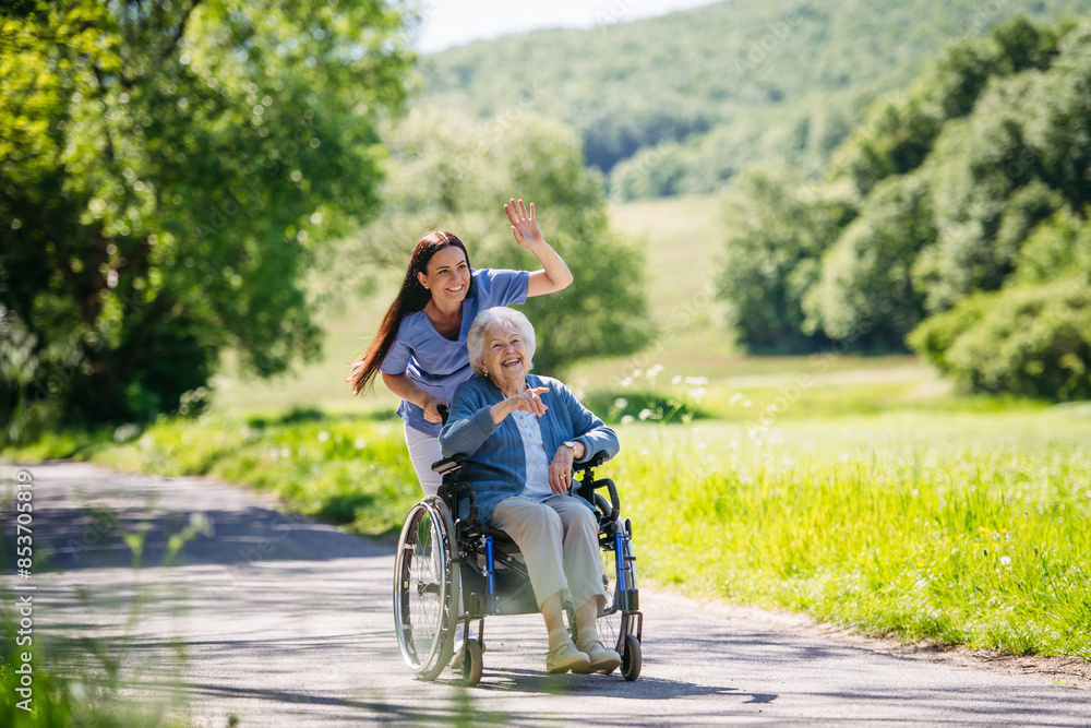Female caregiver pushing senior woman in wheelchair. Nurse and elderly woman enjoying a warm day in nursing home, public park. Female caregiver pushing senior woman in wheelchair. Nurse and elderly woman enjoying a warm day in nursing home, public park.