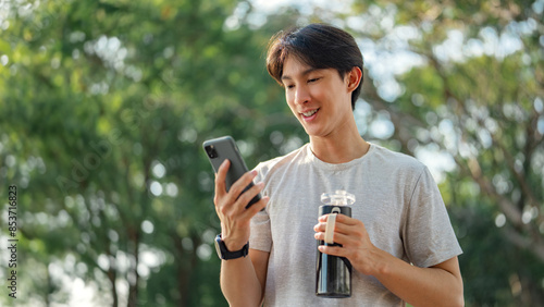 Happy asian man holding mobile phone and bottle of water at public park, Male using smartphone, checking mobile fitness application, copy space.