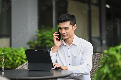 Handsome male entrepreneur having phone conversation and using digital tablet at outdoors cafe