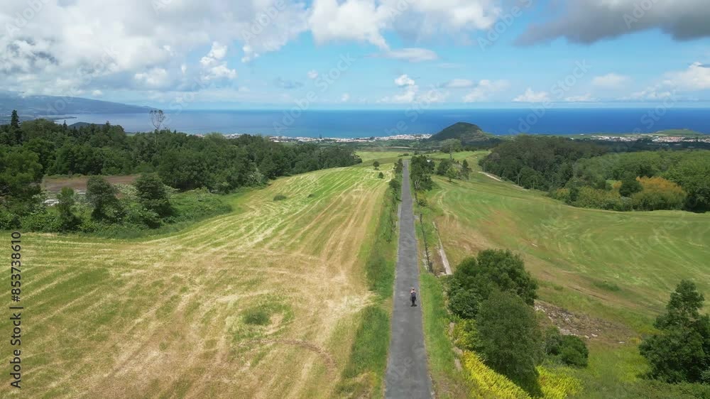 A scenic road stretches through green fields with a distant view of Lagoa do Fogo and the sea A scenic road stretches through green fields with a distant view of Lagoa do Fogo and the sea