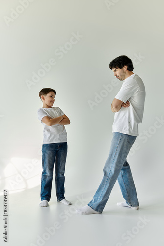 A father and son stand together in a white room, both wearing white t-shirts and jeans.