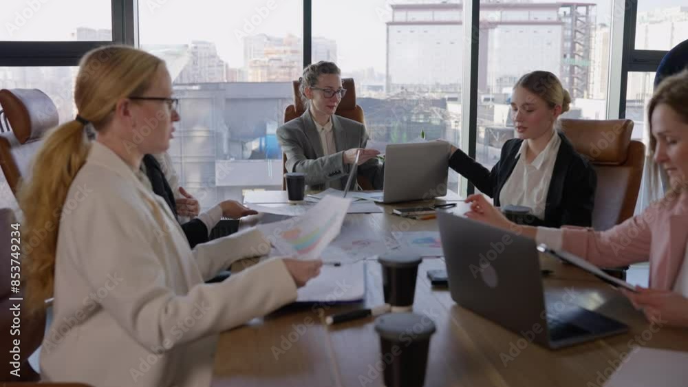 A group of businesswomen in business clothes are communicating during a meeting and passing each other sheets with graphs and ideas about their future work in a modern society with large windows A group of businesswomen in business clothes are communicating during a meeting and passing each other sheets with graphs and ideas about their future work in a modern society with large windows