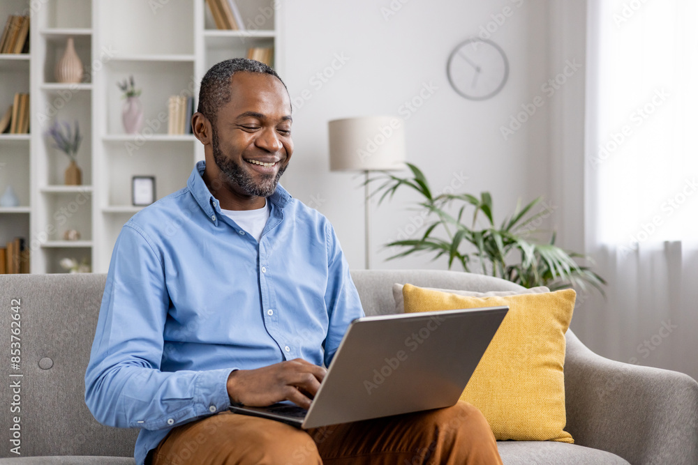 Smiling man working on laptop while sitting on a couch in home office setting with modern decor. Smiling man working on laptop while sitting on a couch in home office setting with modern decor.