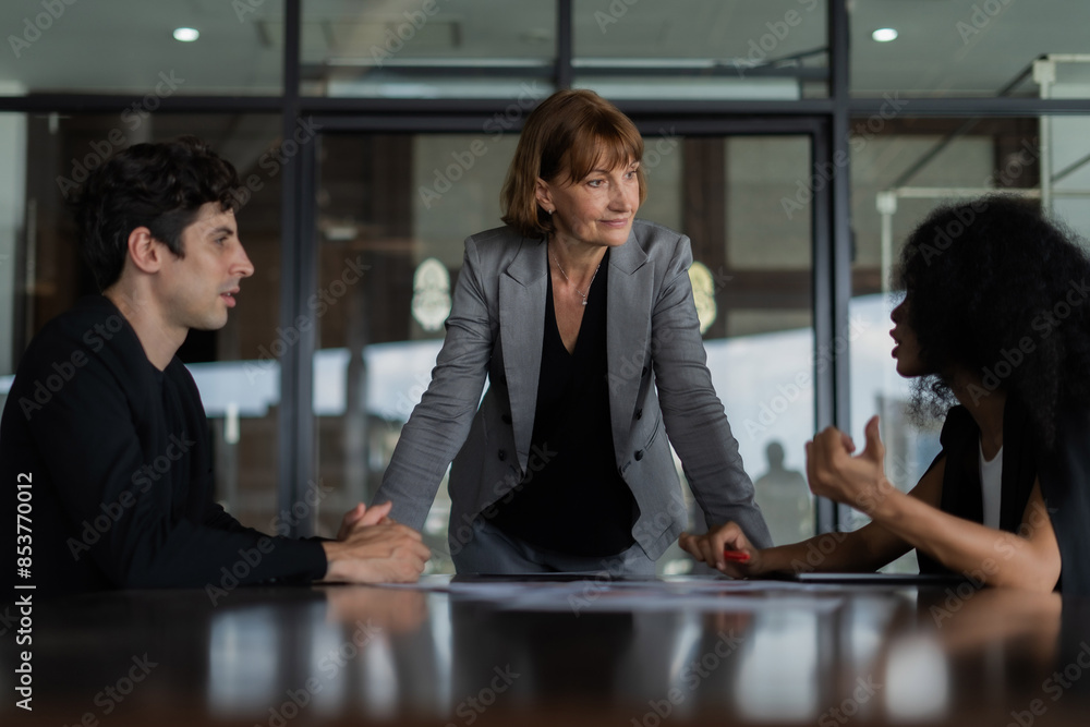 A woman in a suit stood in front of the other two. One of which is a man. A woman is looking at a woman and another person is looking at her. This scene appears to be a business meeting. A woman in a suit stood in front of the other two. One of which is a man. A woman is looking at a woman and another person is looking at her. This scene appears to be a business meeting.