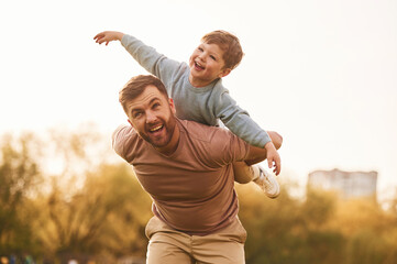 Conception of imagination, sitting on shoulders and flying. Happy father with son are having fun on the field at summertime