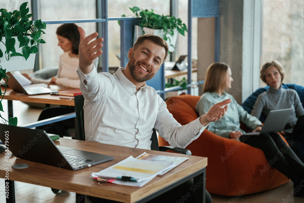 Cheerful friendly man is sitting by the table with laptop. People are working in the office with bean bags chairs in it Cheerful friendly man is sitting by the table with laptop. People are working in the office with bean bags chairs in it