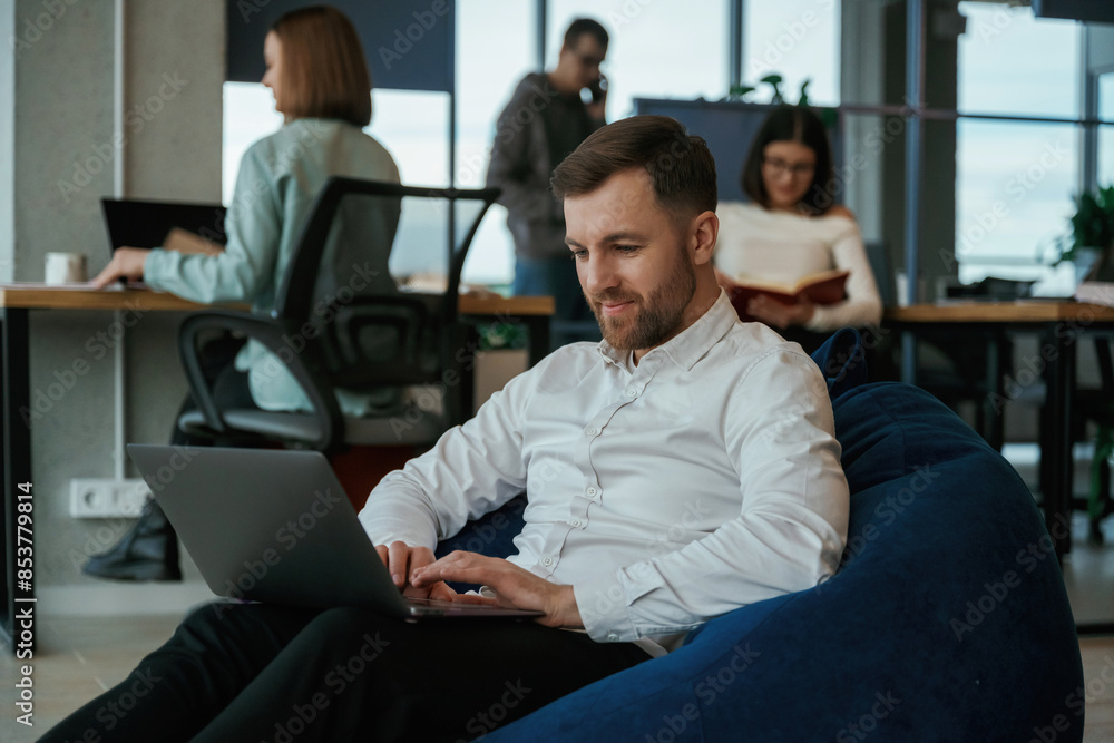 In the blue colored bean bag. With laptop. People are working in the office In the blue colored bean bag. With laptop. People are working in the office