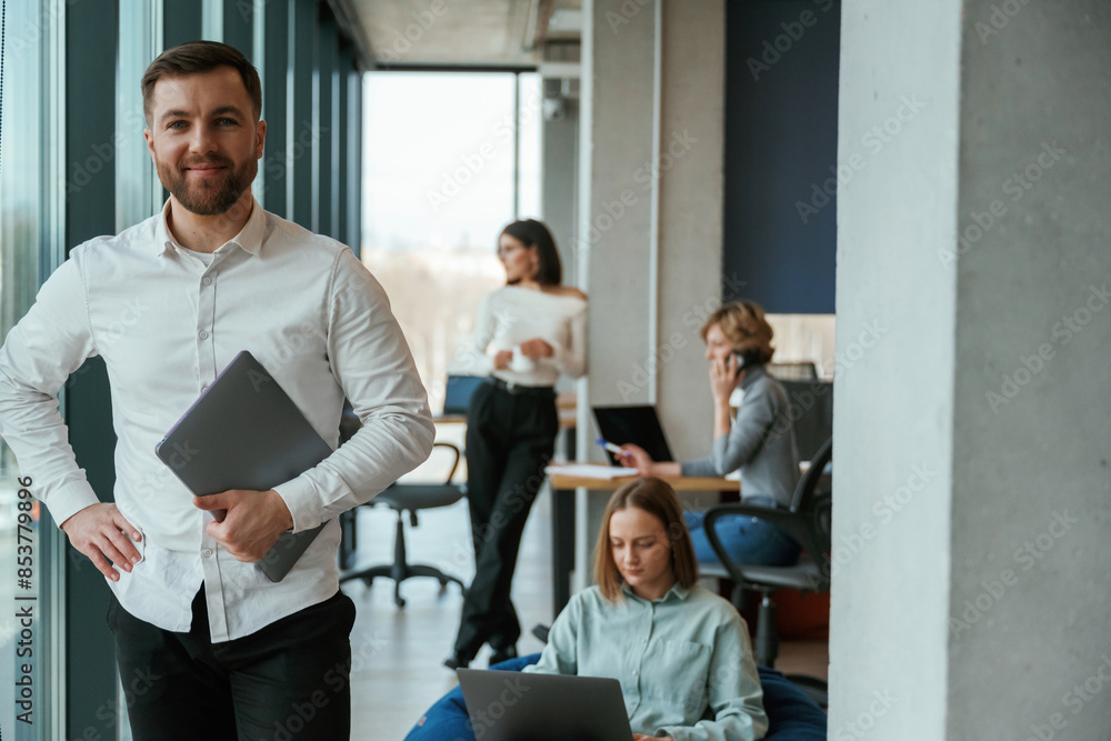 Man is standing and holding laptop. People are working in the office with bean bags chairs in it Man is standing and holding laptop. People are working in the office with bean bags chairs in it