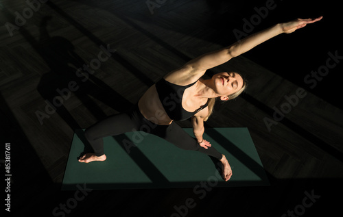 Peaceful young caucasian woman yoga instructor wearing workout clothes practicing vinyasa flow, reverse warrior pose yoga exercise indoors, facing the sunlight with closed eyes. Copy space.