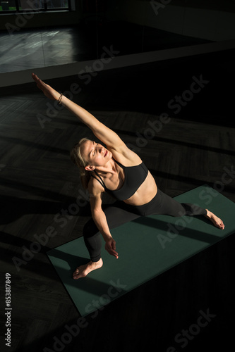 Peaceful young caucasian woman yoga instructor is practicing vinyasa flow, extended side angle yoga ashtanga asana indoors, facing the sunlight with closed eyes. Wellness, lifestyle, copy space. photo