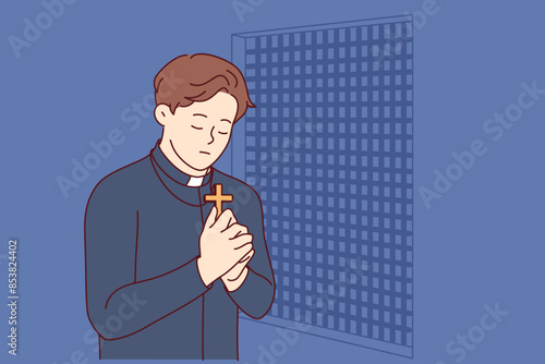 Padre prays and holds christian cross in hands, standing in confession room, asking god to atone for sins of temple visitors. Catholic priest observes ritual of confession and reads prayer