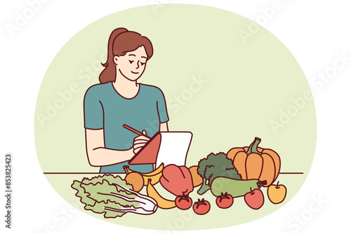 Woman nutritionist is standing near table with vegetables and making notes in notebook, making plan for new diet to detoxify body. Girl nutrition specialist uses checklist to mark diet ingredients