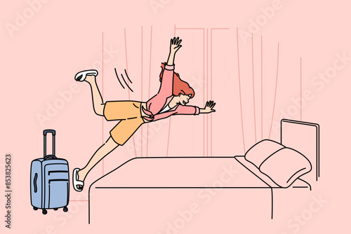 Happy woman tourist jumps on hotel bed, rejoicing at onset of vacation and opportunity to visit sunny resort. Girl with travel suitcase is in hotel and happily plops down on bunk to relax