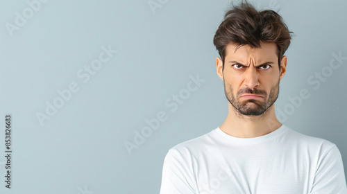 Young man with a displeased expression in white t-shirt, frowning against a plain light blue background, expressing discontent or frustration.