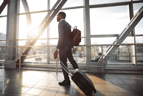 Black businessman is pulling his suitcase at the exterior of a modern airport. The scene is lively and professional, reflecting the efficiency and hustle of business travel.