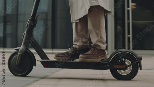 Tracking low section shot of unrecognizable man riding e-scooter on pavement along modern glass office building