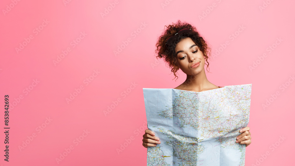 Fototapeta premium A young African American woman with curly hair stands against a soft pink backdrop, carefully examining a large unfolded map. She appears to be planning a journey, copy space