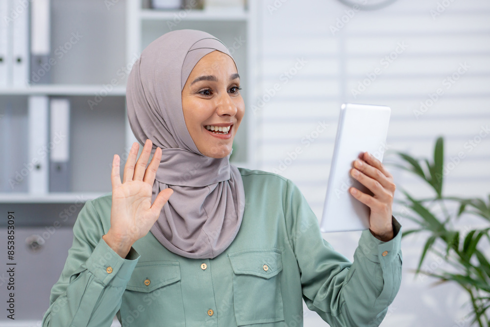 Smiling woman in hijab waving at a tablet during a video call while sitting at home. Connecting with friends or family remotely. Smiling woman in hijab waving at a tablet during a video call while sitting at home. Connecting with friends or family remotely.