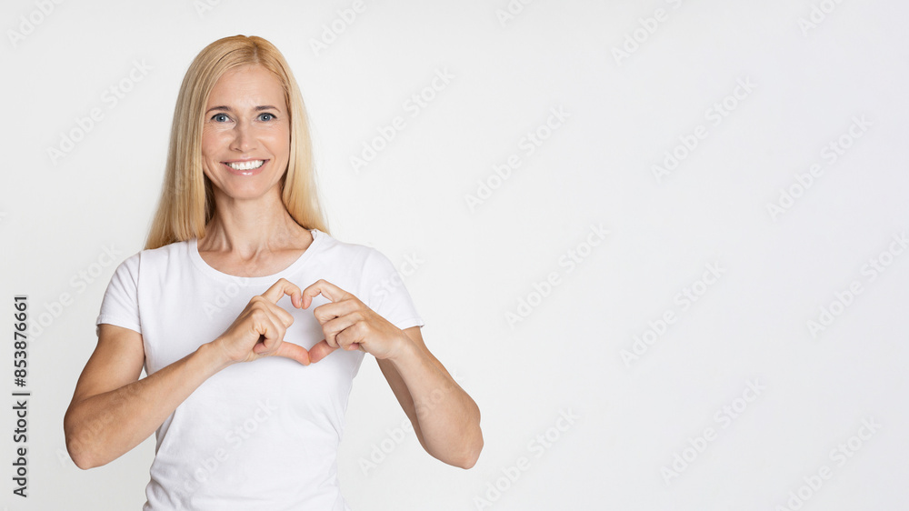 A young woman with blonde hair is standing in front of a white background. She is wearing a white t-shirt and smiling. She is using her hands to make a heart shape in front of her chest. A young woman with blonde hair is standing in front of a white background. She is wearing a white t-shirt and smiling. She is using her hands to make a heart shape in front of her chest.