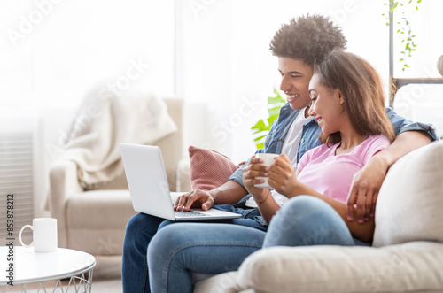African American couple sits on a white couch in a living room, using a laptop and drinking coffee. The guy is sitting behind the girl with his arm around her, and she is looking at the laptop.