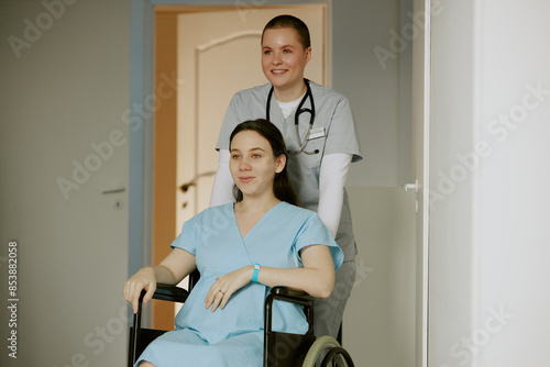 Happy nurse taking her pregnant patient to consulting room while she sitting in wheelchair and resting photo