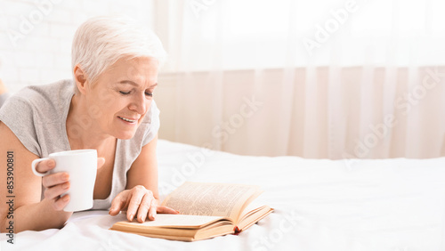 Senior woman is laying on a bed, absorbed in reading a book while sipping a cup of coffee. She appears relaxed and comfortable in her surroundings, copy space