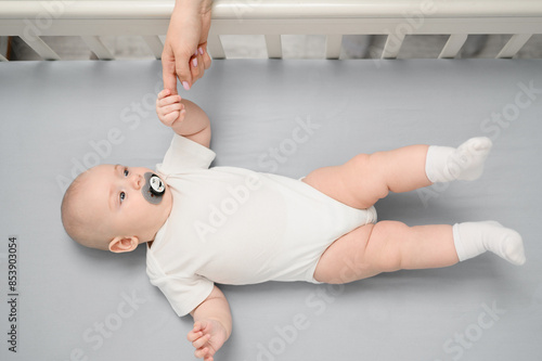 Newborn baby with pacifier in mouth rests peacefully in crib, surrounded by cozy atmosphere of nursery and holds his mom hand, top view. Concept of happiness of motherhood, babyhood, maternity leave